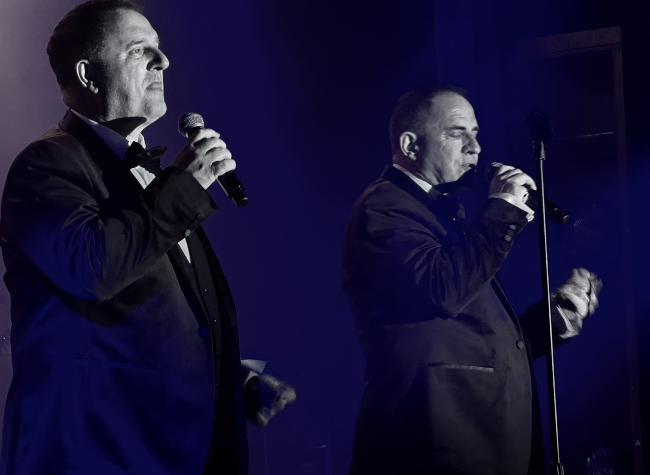 Two performers in tuxedos singing on stage with blue lighting